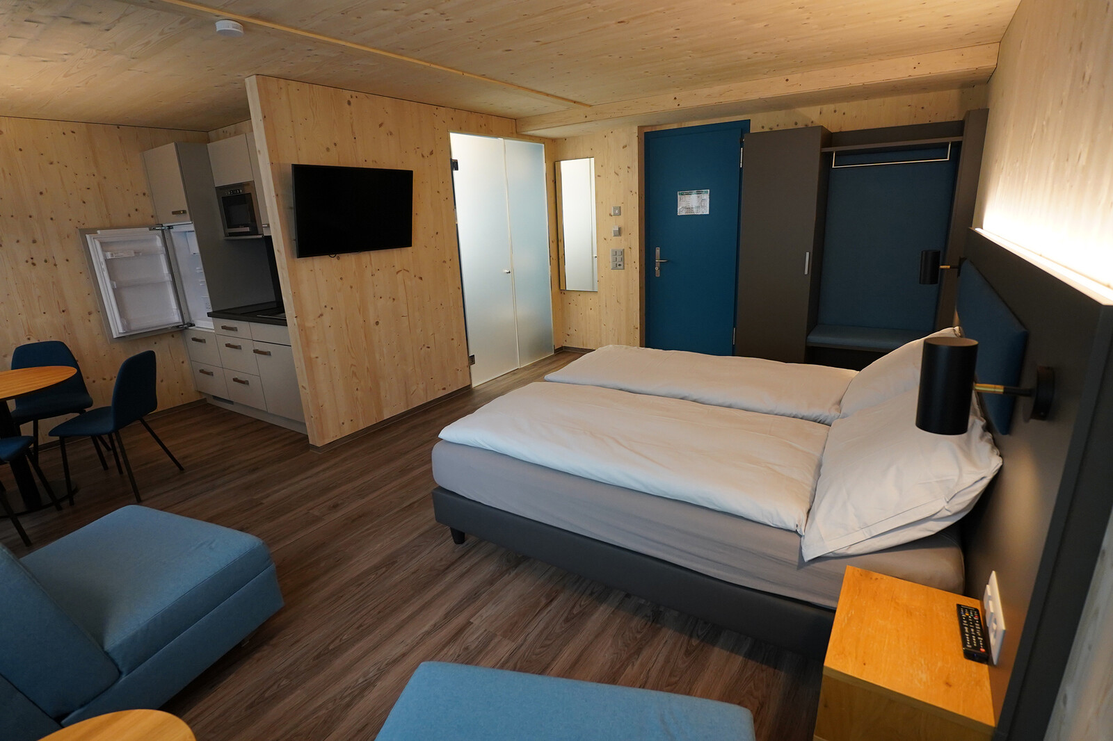 Modern hotel room with light wood walls, a cozy double bed, blue accent chairs, a small kitchen area, mounted TV, and soft ambient lighting.