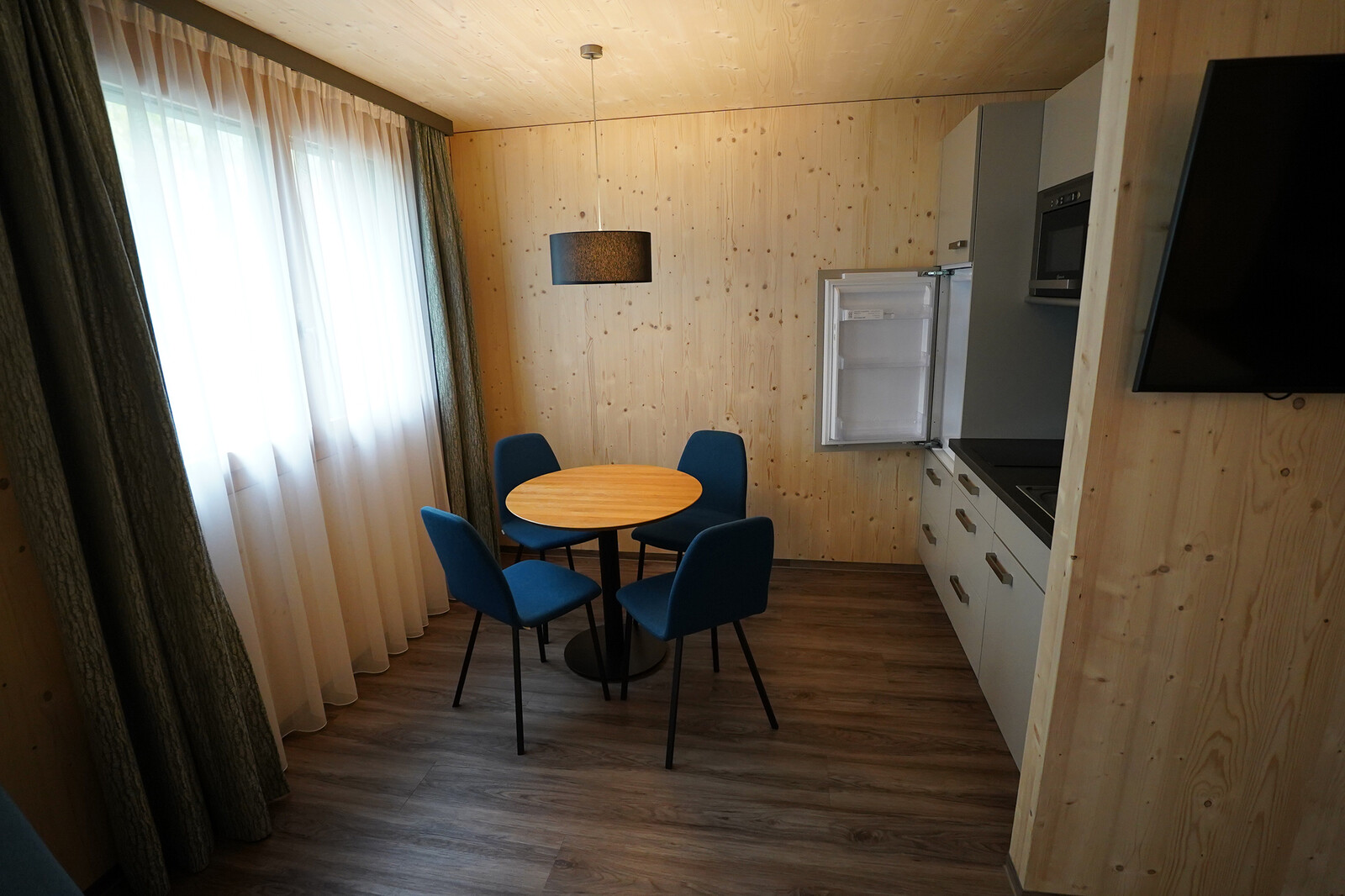 Cozy dining area with a round table and four blue chairs, set against wooden walls. A window with sheer curtains adds soft light. Nearby, an open fridge in a compact kitchenette.
