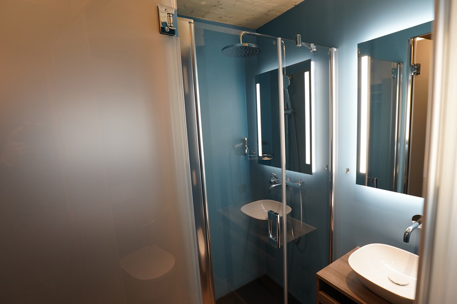 Modern bathroom with a blue wall, shower area with glass doors, and a large illuminated mirror. A sleek white basin sits on a wooden counter, exuding a clean, contemporary feel.