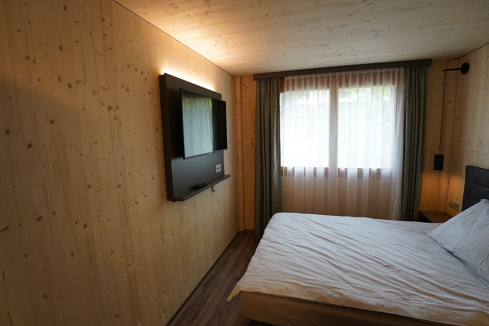 Bedroom with wooden walls and floor, featuring a mounted TV, white bed linens, and a window with sheer curtains. The ambiance is warm and cozy.