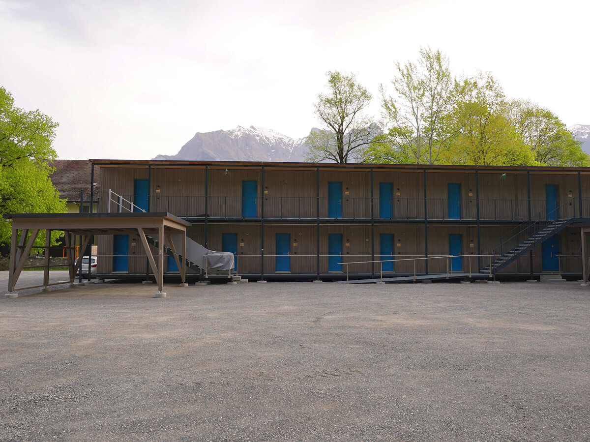Two-story building with wooden facade and bright blue doors, set against a backdrop of trees and snowy mountains. The sky is overcast, creating a serene atmosphere.